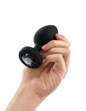 A hand is shown holding the b-Vibe Vibrating Jewel Butt Plug in Black against a blank background on its side, displaying the black crystal on the base.