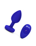The b-Vibe Vibrating Jewel Butt Plug is shown against a blank background next to its 4 buttoned remote. The plug is a size L/XL, which is Blue Sapphire colored.