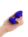 A hand is shown holding the b-Vibe Vibrating Jewel Butt Plug in Blue Sapphire against a blank background on its side, displaying the blue crystal on the base.