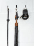 A close-up image of the Sissoo Rosewood Short Handle Leather Flogger with leather hanging strap on a white background. The flogger is shown hanging on a black hook next to leather wrist cuffs and a black silicone ball gag made by The Stockroom.
