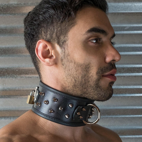 A brunette man with facial stubble is shown from the shoulders up in front of a metal wall. He wears the black leather Alpha Dog Collar with Spikes from the Stockroom, which is locked shut with a brass padlock.