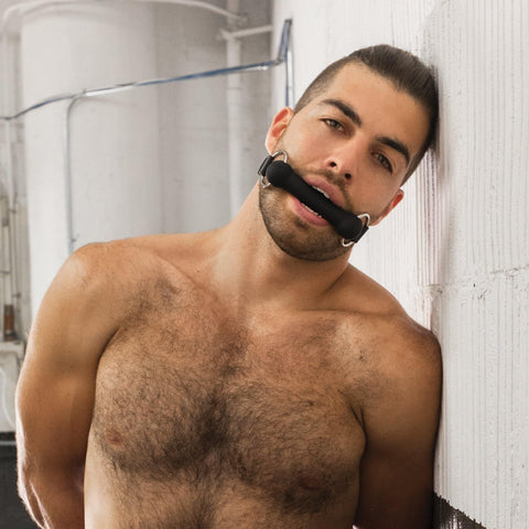 A shirtless brunette man with pulled-back hair and facial stubble is shown from the chest up, leaning his head against a white wall. In his mouth is a matte black silicone bit gag from the Stockroom.
