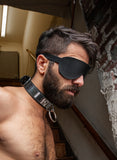 A shirtless male is standing at the bottom of a stairwell wearing the black fleece-lined-leather-blindfold.