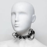 White, faceless mannequin bust wearing a black latex collar with the band woven in and out of several O-rings. There is one large ring on the left side of the neck.