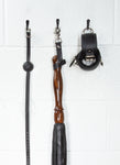A close-up image of the Sissoo Rosewood Long Handle Leather Flogger with leather hanging strap on a white background. The flogger is shown hanging on a black hook next to leather wrist cuffs and a black silicone ball gag made by The Stockroom.
